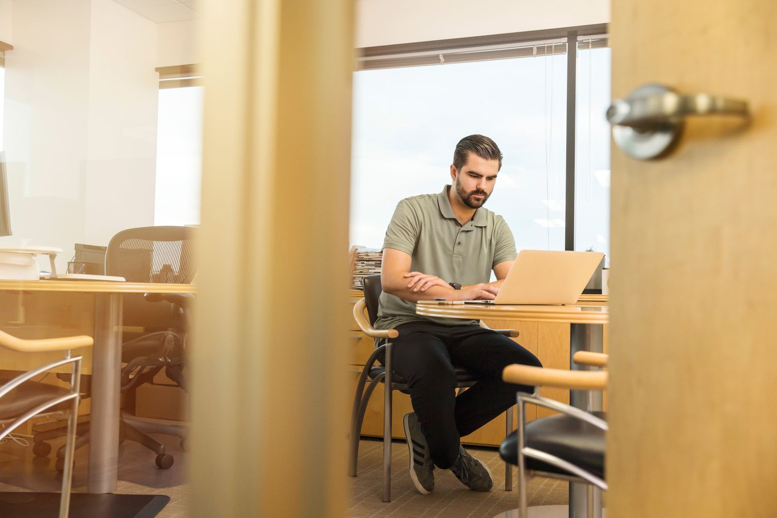 Homem trabalha em um notebook em uma sala de escritório vista através de uma porta entreaberta.