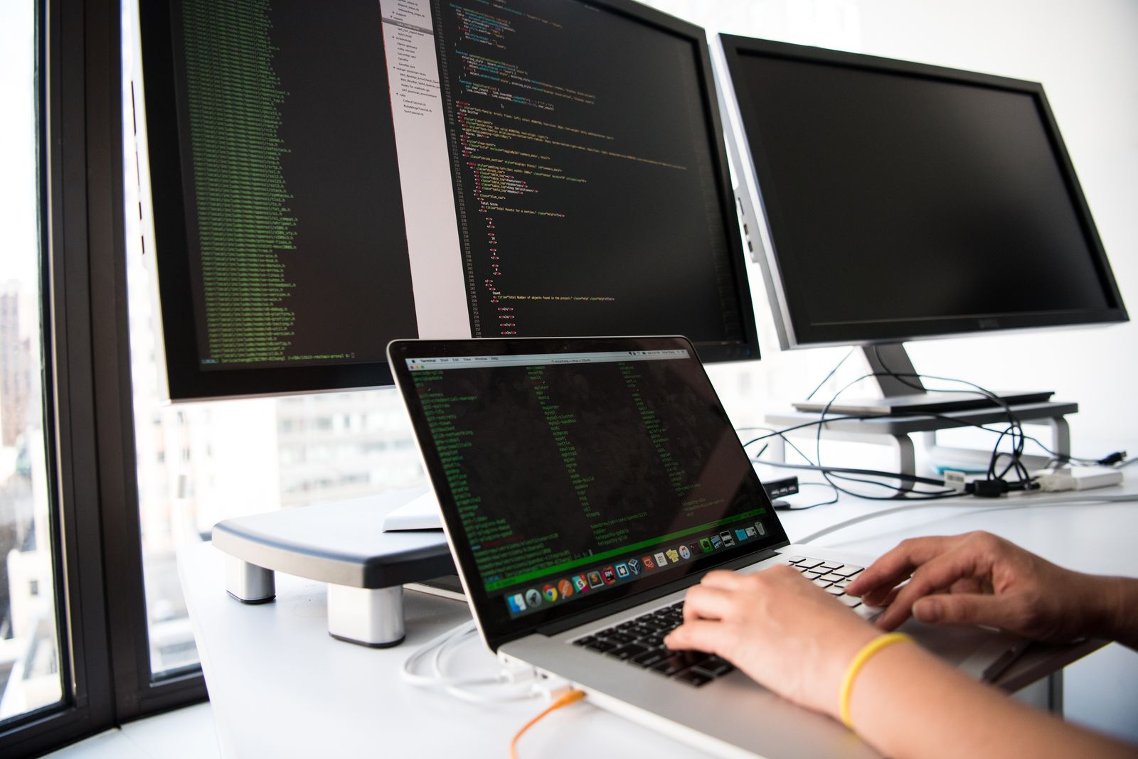 Estação de trabalho com dois monitores e notebook exibindo linhas de código.